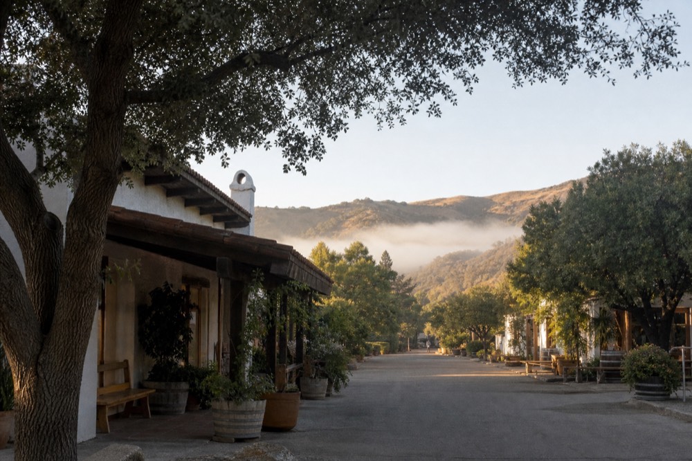 A quiet village street lined with low Spanish-colonial adobe and wood-front tasting rooms shaded by trees, no text, no logos, no people