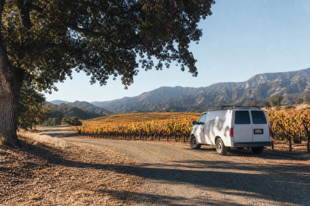 A small touring van parked beside oak-shaded vineyard rows on a quiet ranch road in warm afternoon light, no text, no logos, no people
