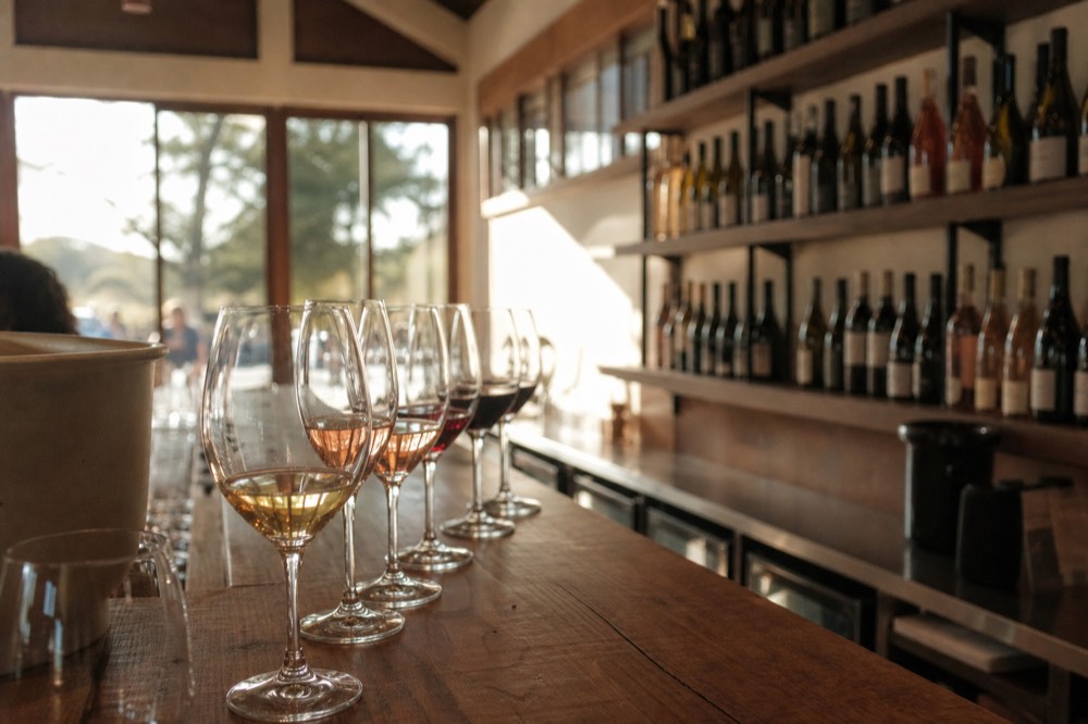 A row of wine glasses on a wooden bar inside a bright Solvang tasting room with bottles lined on shelves behind, no text, no logos, no people