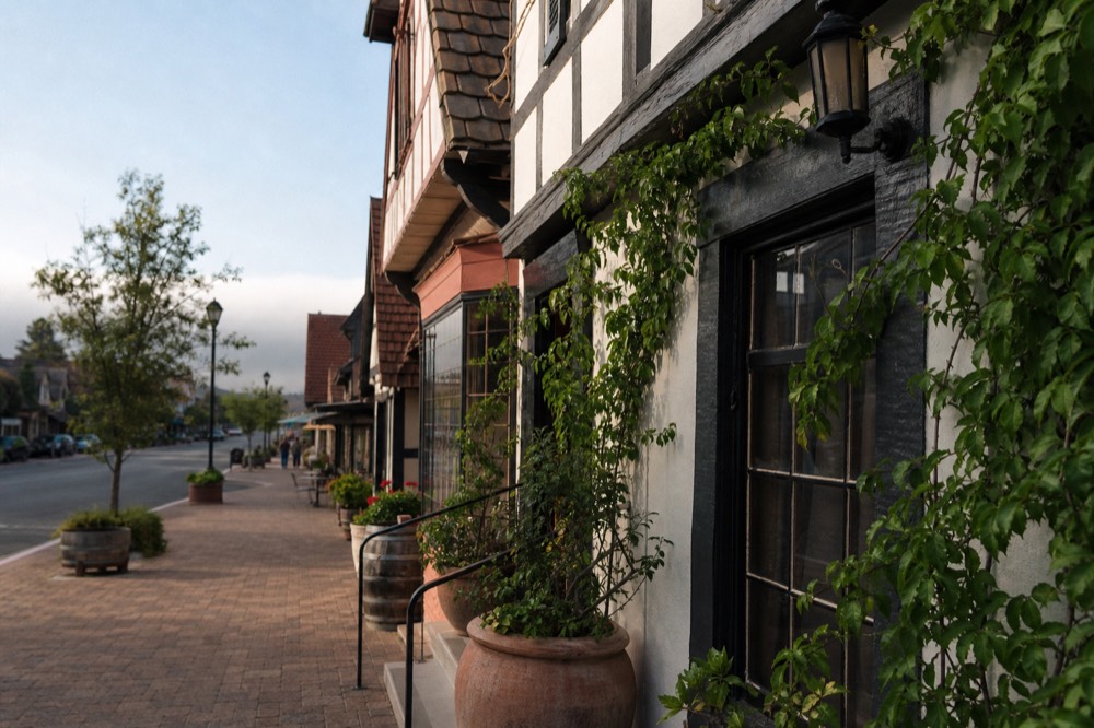 A sunlit Solvang side street lined with small tasting-room storefronts and potted vines, half-timbered rooflines above, no text, no logos, no people
