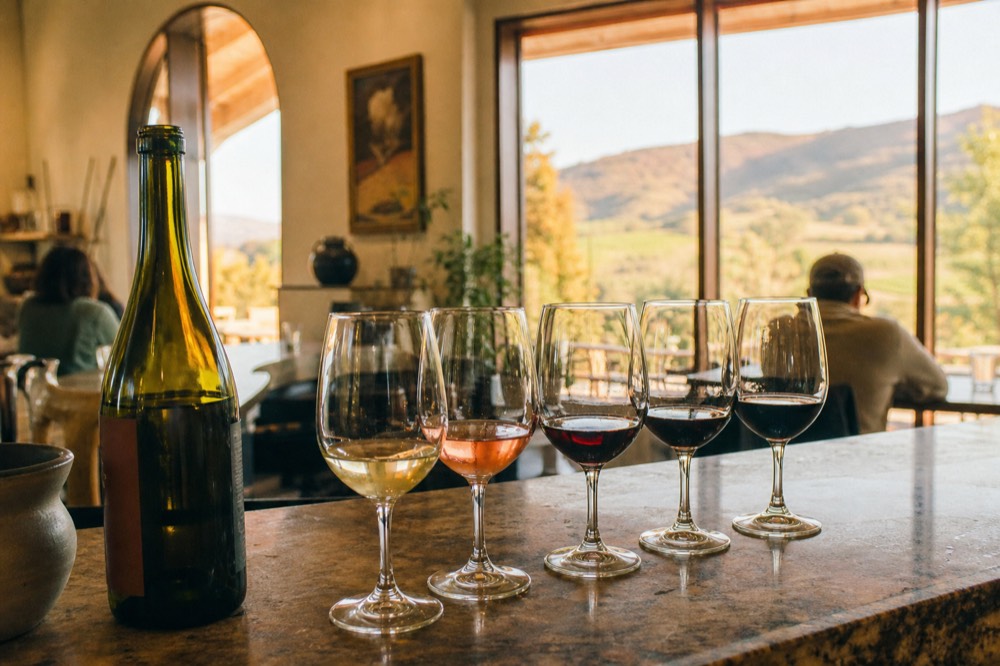 A full tasting flight of glasses set down on a Solvang bar beside an open bottle in a bright interior, no text, no logos, no people