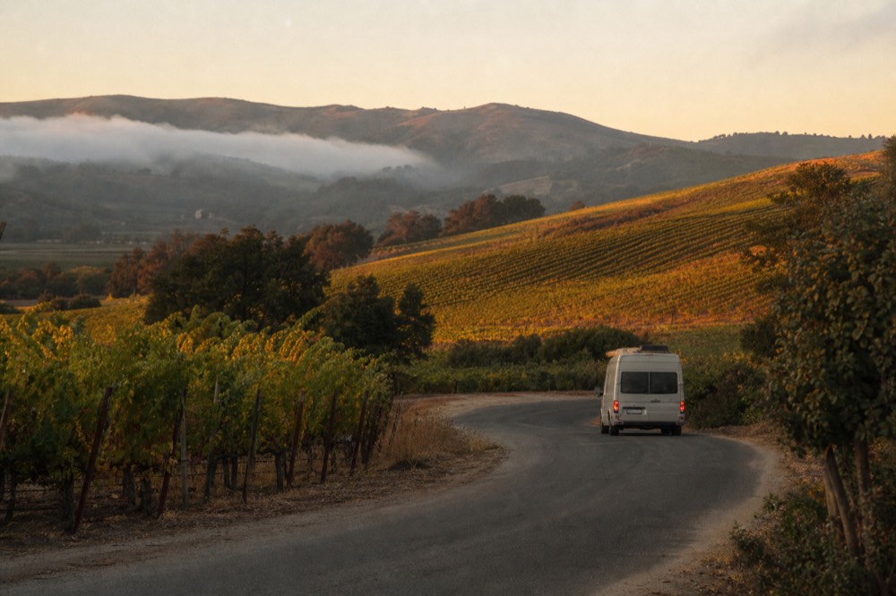 A small touring van on a rural valley road winding between vineyard blocks and low fog-draped hills, no text, no logos, no people